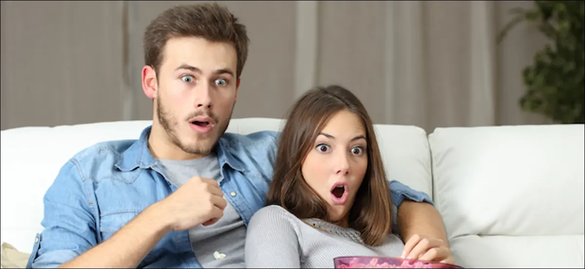 Pareja sorprendida viendo un programa de televisión de realidad sentado en un sofá en casa