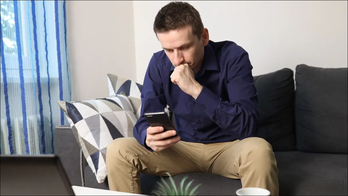 Un jeune homme assis sur un canapé et regardant son téléphone avec une expression inquiète.