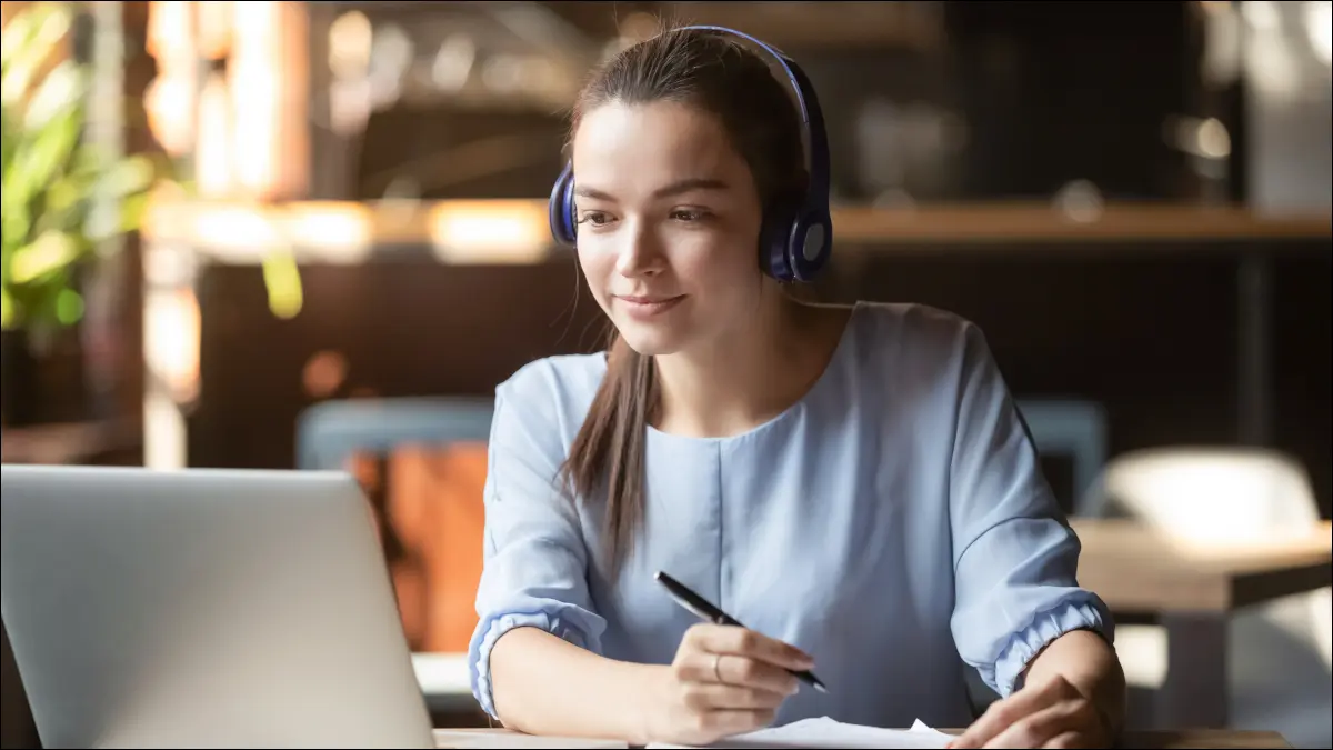 Jeune femme portant des écouteurs tout en prenant des notes devant un ordinateur portable,