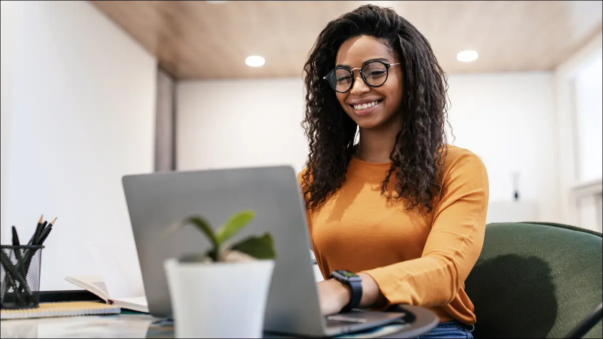 Mujer joven con gafas y sonriendo mientras escribe en una computadora portátil.