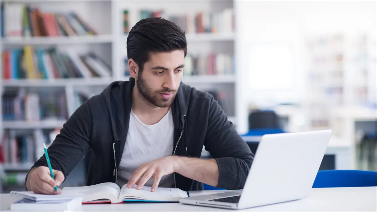 Junger Mann studiert in einer Bibliothek und benutzt einen Laptop.