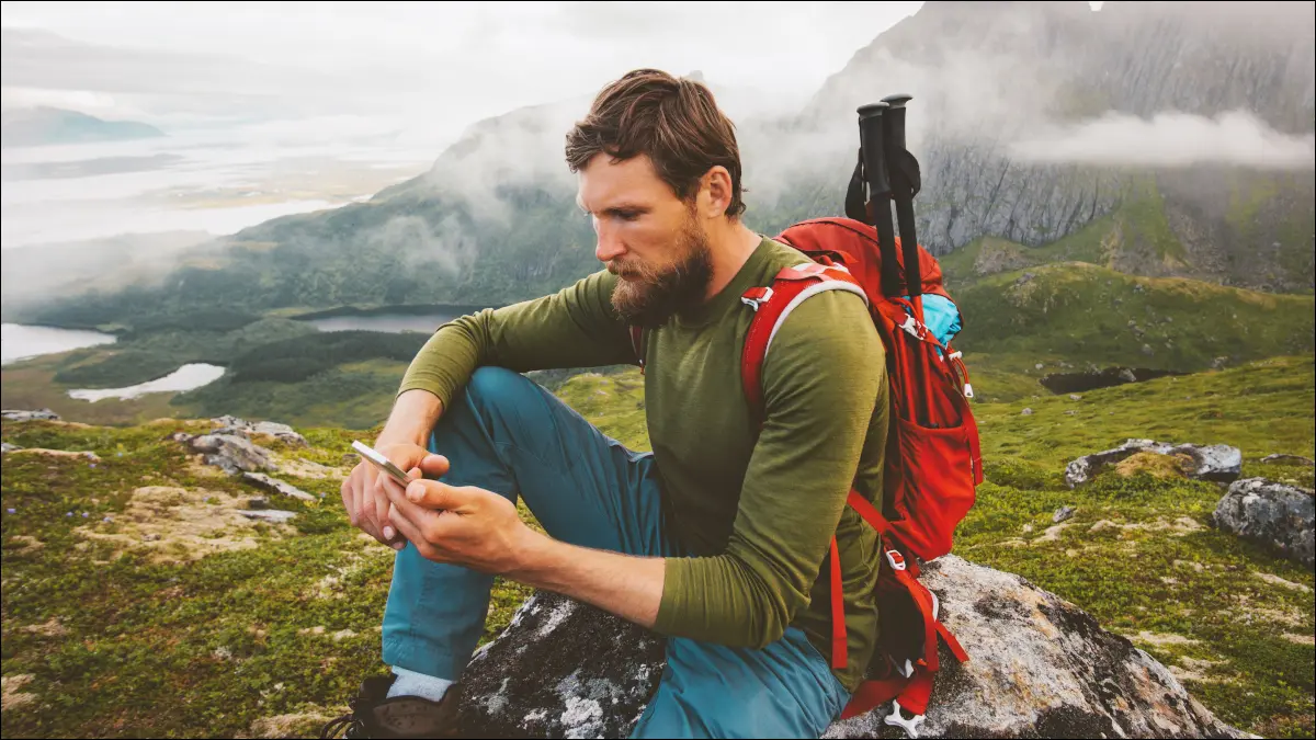 Mann, der einen Rucksack trägt und ein Smartphone auf einem Berggipfel benutzt.