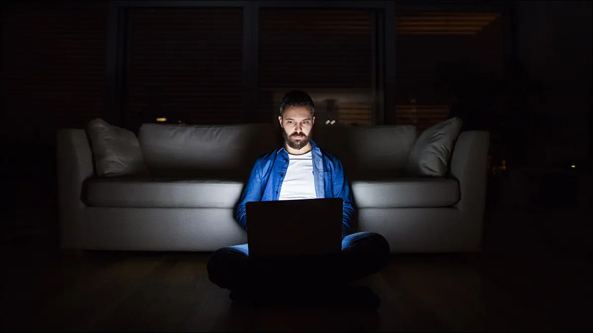 Une personne assise dans une pièce sombre, le visage éclairé par un écran d'ordinateur portable lumineux.