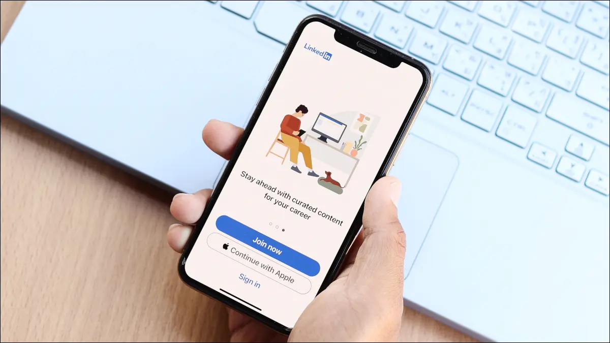 Mano di una persona che tiene uno smartphone sulla tastiera di un laptop, con l'app LinkedIn in mostra.