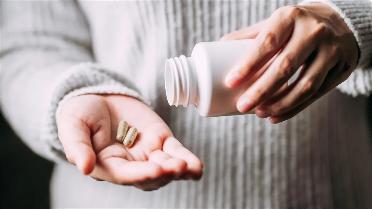 As mãos da pessoa segurando pílulas de ervas medicinais e uma garrafa de plástico branca.