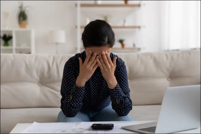 Femme pressant les mains contre son front sous l'effet du stress, avec une calculatrice et un ordinateur portable devant elle.