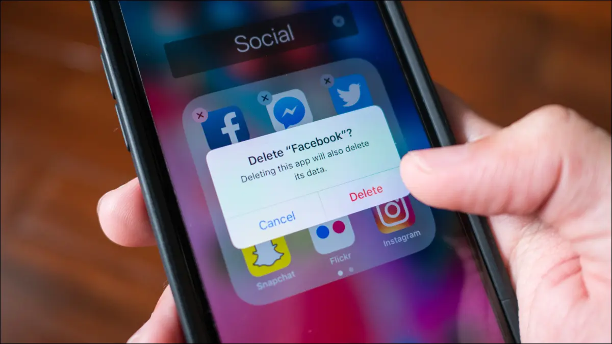 Tangan seseorang memegang iPhone dengan kotak dialog untuk menghapus aplikasi Facebook yang terlihat di layar.