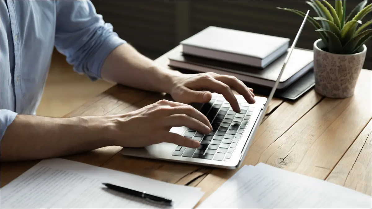 Manos de la persona escribiendo en una computadora portátil con papeleo y cuadernos en el escritorio.